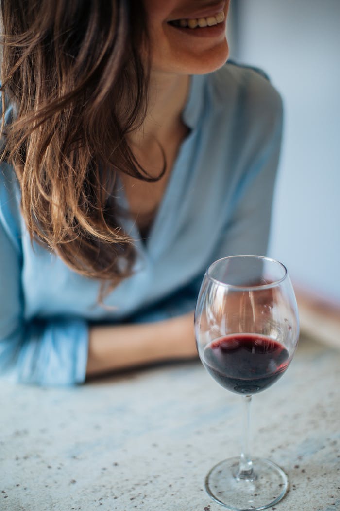 A woman in a blue shirt smiling and enjoying a glass of red wine indoors, conveying relaxation and leisure.